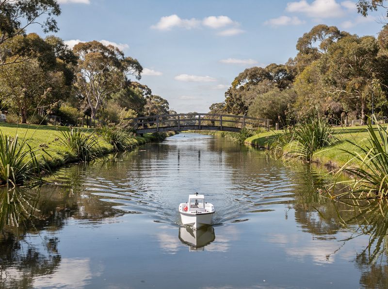 A small white remote-controlled boat on a park canal with flax on the banks and a timber footbridge