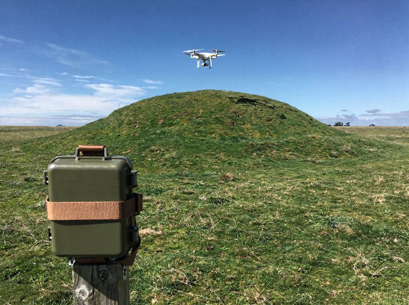 A trail camera on a post facing a rounded grassy hill, with a small drone hovering above the hill