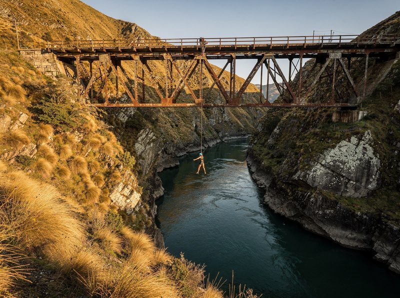 A bungy jumper hanging on the cord below an old steel truss bridge, above a dark green river in a gorge with golden tussock on its slopes