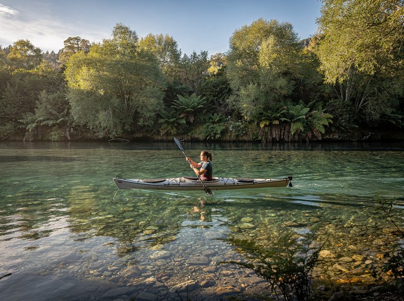 A woman paddling a white kayak along a clear river lined with native bush and tree ferns