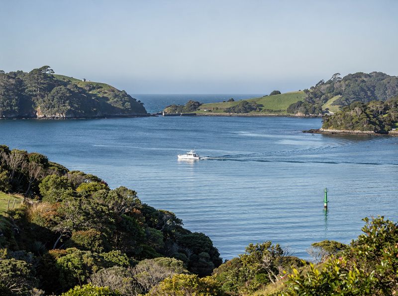 A small boat crossing a calm, bush-clad harbour near a green channel marker