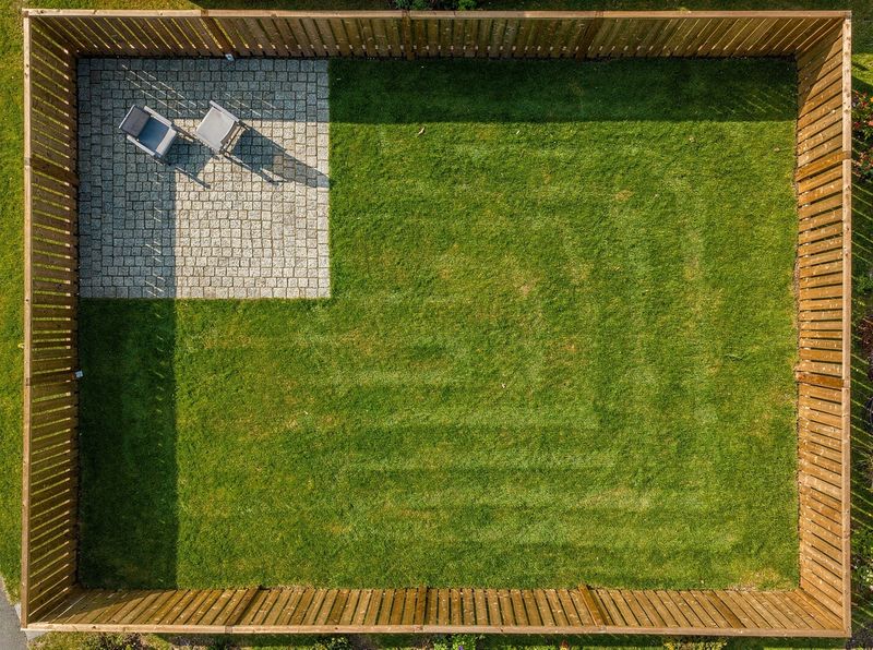 A fenced back lawn seen from above, with a square paved patio in one corner